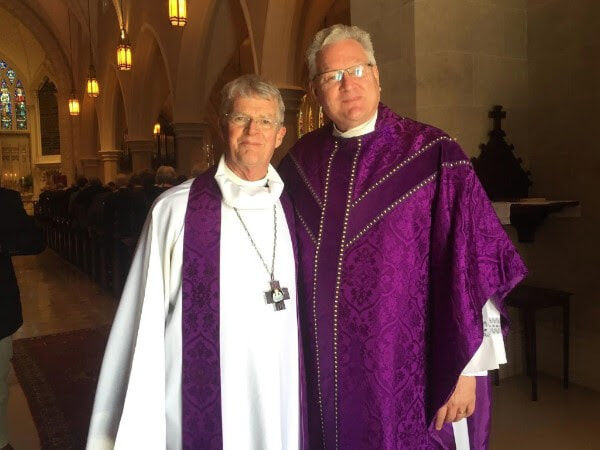 Two men, one wearing a white alb, purple stole, and silver cross on a chain. The other is wearing a purple chasuble.